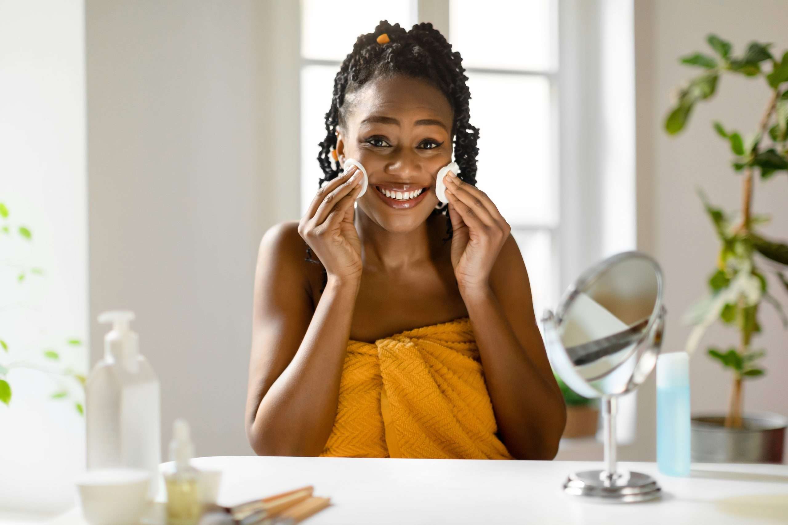 Smiling woman in a yellow towel using cotton pads to cleanse her face in a bright, modern bathroom.